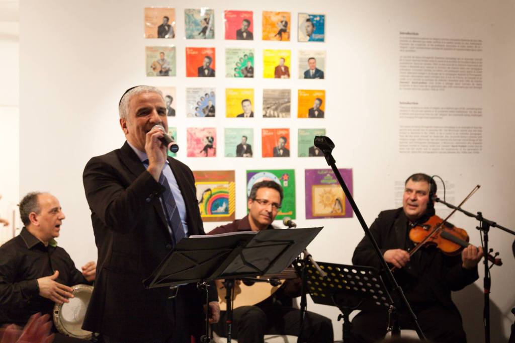 Samy Elmaghribi exhibition. Man sings with musicians behind him. Background display of colourful 45 RPM record covers.